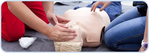 Two people performing chest compressions on a CPR training manikin lying on the floor.