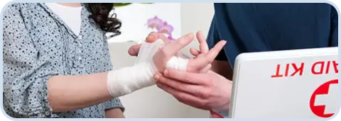 Person with a bandaged hand receives care from another person, in front of a first aid kit with a red cross emblem visible.